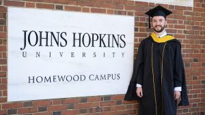 Ryan Demo stands next to the Johns Hopkins University Homewood campus sign in graduation garb.